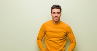 Genç Arap adam gururlu, kendinden emin, havalı, küstah ve küstah görünüyor, gülümsüyor, düz duvara karşı başarılı hissediyor.