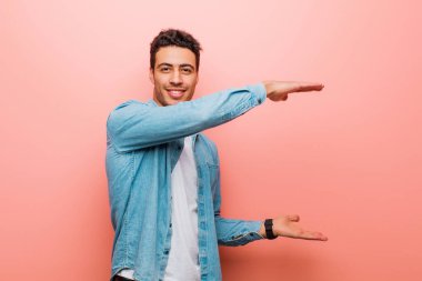 Genç Arap adam gülümsüyor, mutlu, pozitif ve tatmin olmuş hissediyor, fotokopi alanında pembe duvara karşı nesne ya da konsept tutuyor ya da gösteriyor