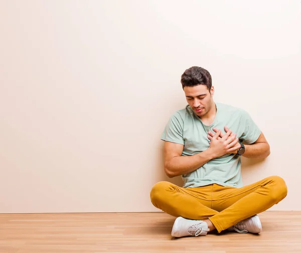 young arabian man looking sad, hurt and heartbroken, holding both hands close to heart, crying and feeling depressed sitting on the floor