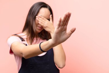 Genç ve güzel bir kadın yüzünü elleriyle kapatıyor ve kamerayı durdurmak için diğer elini öne koyuyor. Pembe duvara karşı fotoğraf ya da resim çekmeyi reddediyor.