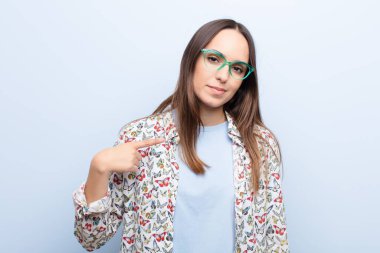 Genç ve güzel bir kadın gururlu, kendinden emin ve mutlu görünüyor, gülümsüyor ve kendini işaret ediyor ya da mavi duvara karşı bir numara işareti yapıyor.