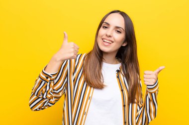 Genç ve güzel bir kadın neşeyle gülümsüyor ve mutlu görünüyor, kaygısız ve pozitif hissediyor, iki başparmağı turuncu duvara dayalı.