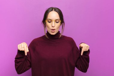 İki eliyle aşağıya bakan ağzı açık genç, güzel bir kadın. Şok olmuş, şaşkın ve şaşkın bir halde mor duvara bakıyor.