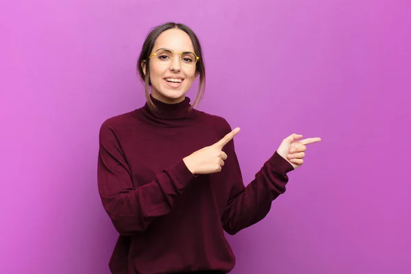 young pretty woman smiling happily and pointing to side and upwards with both hands showing object in copy space against purple wall