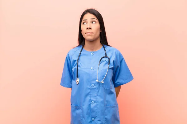 young latin nurse wondering, thinking happy thoughts and ideas, daydreaming, looking to copy space on side against pink wall