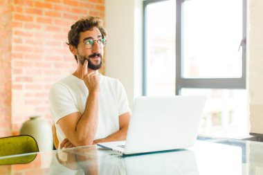 Dizüstü bilgisayarı olan genç sakallı adam mutlu bir şekilde gülümsüyor ve hayal kuruyor ya da şüphe ediyor.