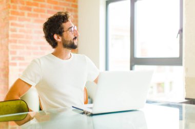 Dizüstü bilgisayarı olan genç sakallı adam mutlu, neşeli ve kendinden emin görünüyor gururla gülümsüyor ve iki elini de kalçasında gezdirmek istiyor.