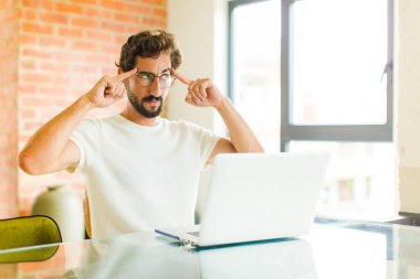 Dizüstü bilgisayarı olan genç sakallı bir adam, kafası karışık ya da şüpheci, bir fikre odaklanmış, çok düşünüyor, kenarda boşluğu kopyalamaya çalışıyor.