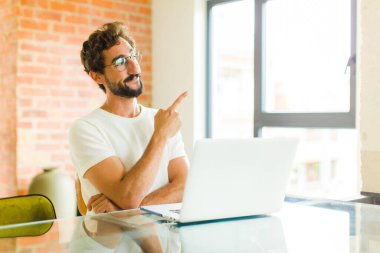 Dizüstü bilgisayarı olan genç sakallı adam mutlu bir şekilde gülümsüyor ve yan bakıyor, merak ediyor, düşünüyor ya da bir fikri var mı?