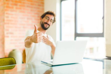 Dizüstü bilgisayarı olan genç sakallı adam gururlu, kaygısız, kendinden emin ve mutlu, başparmağıyla olumlu gülümsüyor.