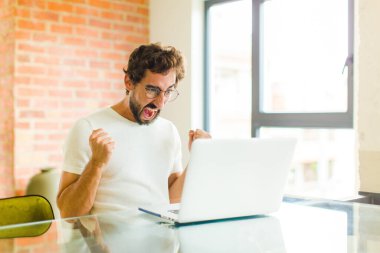 Dizüstü bilgisayarlı genç sakallı bir adam sinirli, sinirli, sinirli bakışlı ve yumruklarını sıkmış, öfkeli bir şekilde bağırıyor.