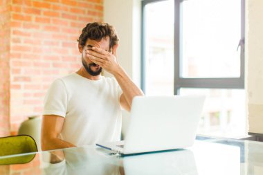 Dizüstü bilgisayarı olan genç sakallı bir adam. Tek eliyle gözlerini kapatıyor. Korkmuş, endişeli, merak ediyor ya da körü körüne bir sürpriz bekliyor.