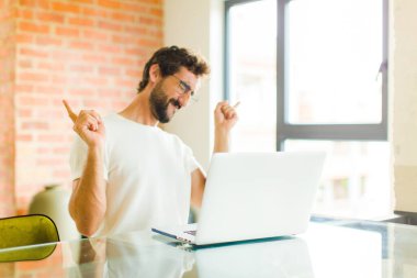 Dizüstü bilgisayarlı genç sakallı adam gülümsüyor, kaygısız hissediyor, rahat ve mutlu, dans ediyor ve müzik dinliyor, bir partide eğleniyor.