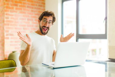 Dizüstü bilgisayarı olan genç sakallı adam mutlu, heyecanlı, şaşırmış ya da şok olmuş hissediyor, gülümsüyor ve inanılmaz bir şeye şaşırıyor.