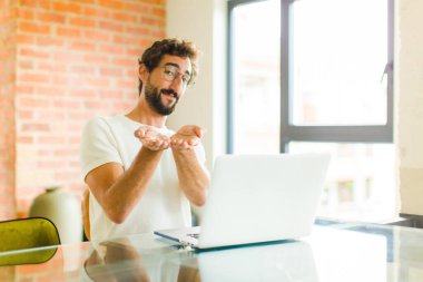 Bilgisayarlı genç sakallı adam mutlu bir şekilde gülümsüyor, kendine güvenen, olumlu bakışlı, bir nesne ya da konsept sunuyor ve gösteriyor.