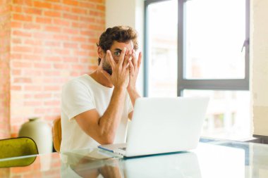 Dizüstü bilgisayarı olan genç sakallı adam şok olmuş, korkmuş ya da korkmuş görünüyor. Yüzü elle kaplanmış ve parmaklarının arasından gizlice bakıyor.