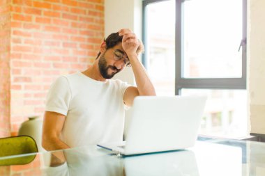 Dizüstü bilgisayarlı genç sakallı adam stresli, yorgun ve hayal kırıklığına uğramış görünüyor alnından ter damlıyor, umutsuz ve bitkin hissediyor.