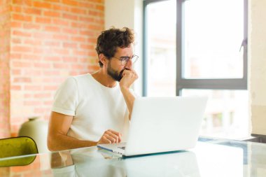 Dizüstü bilgisayarı olan genç sakallı bir adam ciddi, düşünceli ve endişeli, elleri çenesine bastırılmış bir şekilde yan bakıyor.