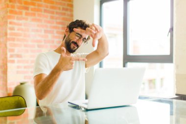 Dizüstü bilgisayarı olan genç sakallı bir adam mutlu, arkadaş canlısı ve pozitif hissediyor, gülümsüyor ve elleriyle bir portre veya fotoğraf çerçevesi yapıyor.