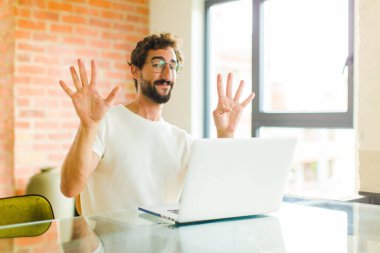 Dizüstü bilgisayarı olan genç sakallı adam gülümsüyor ve dost canlısı görünüyor. 9 ya da 9 numarayı gösteriyor. Eli önde, geriye doğru sayıyor.