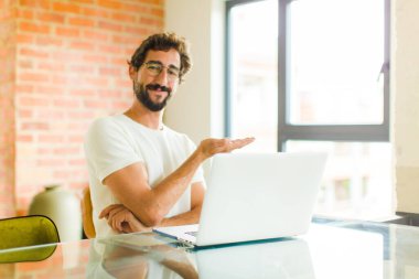 Dizüstü bilgisayarı olan genç sakallı bir adam mutlu bir şekilde gülümsüyor ve fotokopi alanında avuç içi dolu bir konsept gösteriyor.