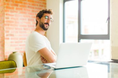 Dizüstü bilgisayarı olan genç sakallı bir adam mutlu, mutlu ve rahat hissediyor, kollarını kavuşturup yan tarafa bakıyor.