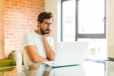 Dizüstü bilgisayarı olan genç sakallı bir adam şok ve inançsızlık içinde, elleri ve yanakları çaprazlanmış, sersemlemiş ve şaşırmış hissediyor.