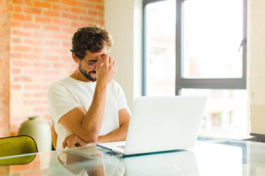 Dizüstü bilgisayarı olan genç sakallı bir adam stresli, utanmış ya da üzgün görünüyor, baş ağrısı var, yüzünü eliyle kapatıyor.