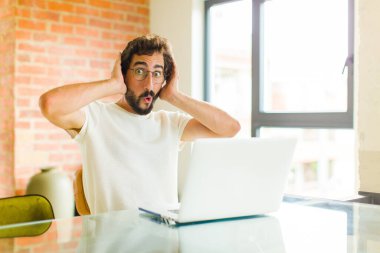 Dizüstü bilgisayarı olan genç sakallı bir adam heyecanlı ve şaşırmış görünüyor, ağzı açık, iki eli başında, kendini şanslı bir kazanan gibi hissediyor.