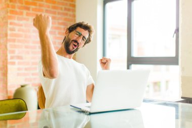 Dizüstü bilgisayarlı genç sakallı adam muzaffer bir şekilde bağırıyor. Heyecanlı, mutlu ve şaşkın bir kazanan gibi görünüyor.