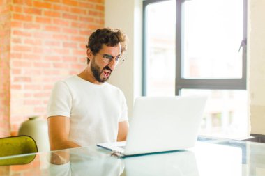 Dizüstü bilgisayarı olan genç sakallı bir adam şok olmuş, kızgın, sinirli ya da hayal kırıklığına uğramış, ağzı açık ve öfkeli görünüyor.