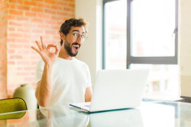 Dizüstü bilgisayarı olan genç sakallı bir adam kendini başarılı ve tatmin olmuş hissediyor ağzı açık gülümsüyor, elle imza atıyor.