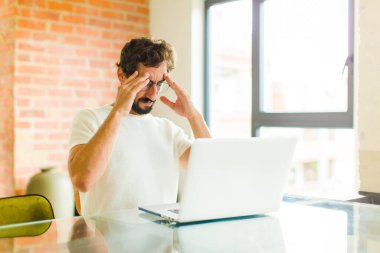 Dizüstü bilgisayarı olan genç sakallı bir adam stresli ve sinirli görünüyor, baskı altında çalışıyor ve başı ağrıyor.