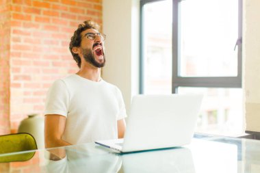 Dizüstü bilgisayarlı genç sakallı adam öfkeyle bağırıyor, agresif bir şekilde bağırıyor, stresli ve sinirli görünüyor.