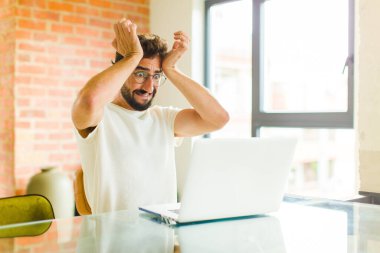 Dizüstü bilgisayarı olan genç sakallı bir adam stresli ve endişeli, depresif ve baş ağrısı yüzünden hayal kırıklığına uğramış, iki elini de başa kaldırıyor.