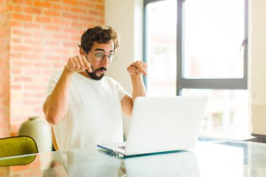 Diz üstü bilgisayarı olan genç sakallı bir adam iki eliyle aşağıya bakıyor, şaşkın, şaşkın ve şaşırmış görünüyor.