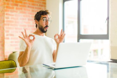 Dizüstü bilgisayarı olan genç sakallı adam şok olmuş, şaşırmış ve şaşırmış hissediyor. İki eliyle de onay imzası atıyor.