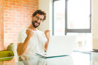 Dizüstü bilgisayarı olan genç sakallı bir adam zaferi kutlarken gülüyor, mutlu ve heyecanlı hissediyor.