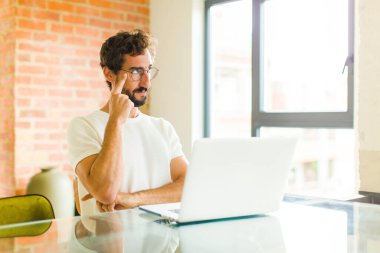 Dizüstü bilgisayarı olan genç sakallı bir adam şüpheli bir ifadeyle yukarı ve yana bakıyor.