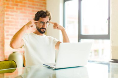 Ciddi ve konsantre görünümlü, beyin fırtınası yapan ve zorlu bir sorunu düşünen, dizüstü bilgisayarlı genç sakallı bir adam.