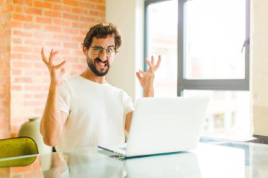 Dizüstü bilgisayarı olan genç sakallı bir adam elleri havada bağırıyor. Öfkeli, sinirli, stresli ve üzgün hissediyor.