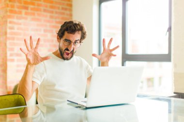 Dizüstü bilgisayarı olan genç sakallı bir adam panik içinde ya da öfkeyle bağırıyor, şok olmuş, korkmuş ya da öfkeli, elleri başının yanında.