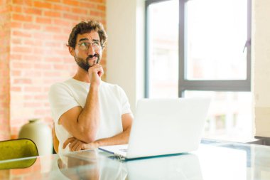 Dizüstü bilgisayarı olan genç sakallı bir adam, şüpheli ve kafası karışık, farklı seçenekleri olan, hangi kararı vereceğini merak eden.