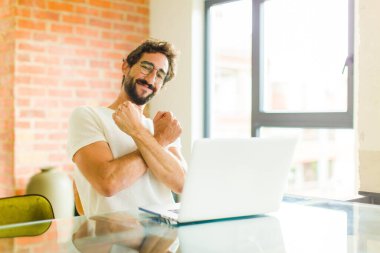 Dizüstü bilgisayarı olan genç sakallı bir adam neşeli bir şekilde gülümsüyor ve kollarını kavuşturup kollarını kavuşturuyor, mutlu ve pozitif hissediyor.