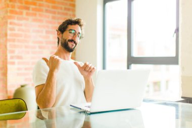 Dizüstü bilgisayarlı genç sakallı adam neşeli bir şekilde gülümsüyor ve yan taraftaki fotokopi alanını gösteriyor, mutlu ve tatmin olmuş hissediyor.