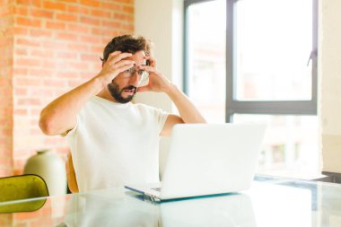 Dizüstü bilgisayarı olan genç sakallı bir adam dehşete kapılmış ve şok olmuş hissediyor, ellerini havaya kaldırıyor ve bir hata yüzünden panikliyor.