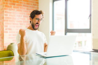 Dizüstü bilgisayarı olan genç sakallı bir adam sinirli bir ifadeyle ya da yumruklarıyla başarıyı kutlarken agresif bir şekilde bağırıyor.