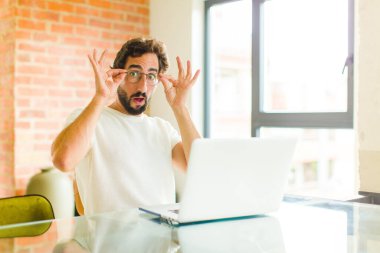 Dizüstü bilgisayarı olan genç sakallı bir adam şok olmuş, şaşırmış ve şaşırmış hissediyor, elinde şaşkın ve inanmayan bir bakışla gözlük tutuyor.