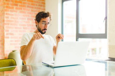 Dizüstü bilgisayarı olan genç sakallı bir adam şok olmuş, ağzı açık ve şaşkın, inançsızlık ve şaşkınlık içinde bakıyor ve işaret ediyor.
