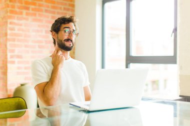 Dizüstü bilgisayarı olan genç sakallı bir adam stresli, hayal kırıklığına uğramış ve yorgun, endişeli ve sorunlu bir bakışla ağrılı boynunu okşuyor.